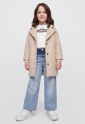 Jeune fille portant un manteau beige, un crop top blanc, un jean bleu clair à jambes larges et des baskets blanches, debout devant un fond clair uni.