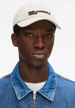 Mladi moški, ki nosi belo kapo z napisom "KARL LAGERFELD JEANS" in modro denim jakno z zadrgo preko bele majice.