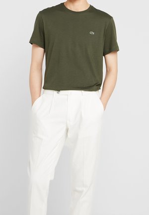 Uomo che indossa una t-shirt verde oliva a maniche corte con piccolo logo e pantaloni bianchi plissettati a vita alta, mani nelle tasche.
