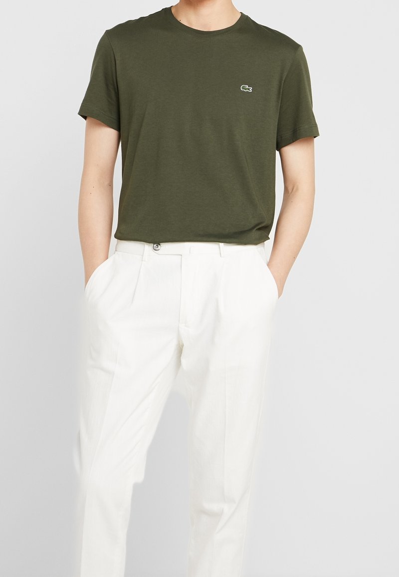 Homme portant un t-shirt vert olive à manches courtes avec un petit logo et un pantalon blanc plissé taille haute, mains dans les poches.