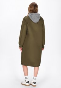 Olíva zöld hosszú puffertuc vékony szürke kapucnival, egyenes szabással, hosszú ujjakkal és bordás mandzsettával, fehér sportcipők felett viselve.