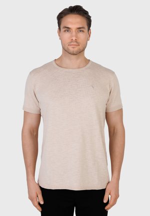 Nuori mies, jolla on lyhyet tummat hiukset, pukeutunut beigen väriseen lyhythihamaiseen paitaan ja mustiin housuihin, seisoo rennosti käsivarret alas suorassa yksiväristä taustaa vasten.