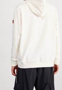 Sudadera blanca con bolsillo canguro, puños y dobladillo acanalados. Presenta un parche de logo en contraste en la manga izquierda. Tejido suave y texturizado.