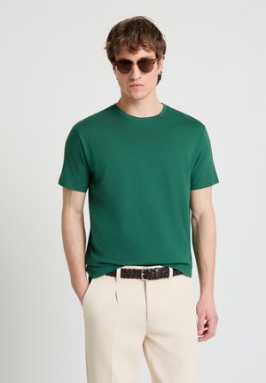 Giovane uomo con occhiali da sole marroni, camicia verde a maniche corte, pantaloni beige e cintura intrecciata scura in piedi con le mani in tasca.