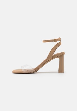 Sandale beige à talon haut avec bride transparente à l'avant, bride à la cheville avec boucle et talon bloc sur fond blanc uni.