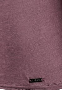 Lila Kurzarmoberteil aus strukturiertem Stoff. Mit genähtem Saum und einem kleinen schwarzen Logotag unten links.