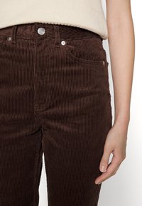 Gros plan sur une personne portant un pantalon en velours côtelé marron foncé avec des poches avant visibles et un bouton en argent, la main reposant sur le côté.