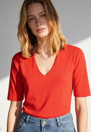 Femme aux cheveux blonds mi-longs portant un haut rouge à col en V et manches courtes, ainsi qu'un jean bleu, sur un fond clair.