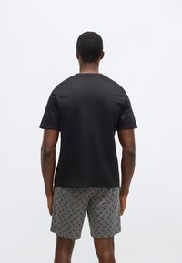 Μαύρο πουκάμισο με κοντά μανίκια και ομαλή υφή, σε συνδυασμό με γκρι σορτς που έχουν επαναλαμβανόμενο σκουρόχρωμο μοτίβο. Βλέπουμε από πίσω.