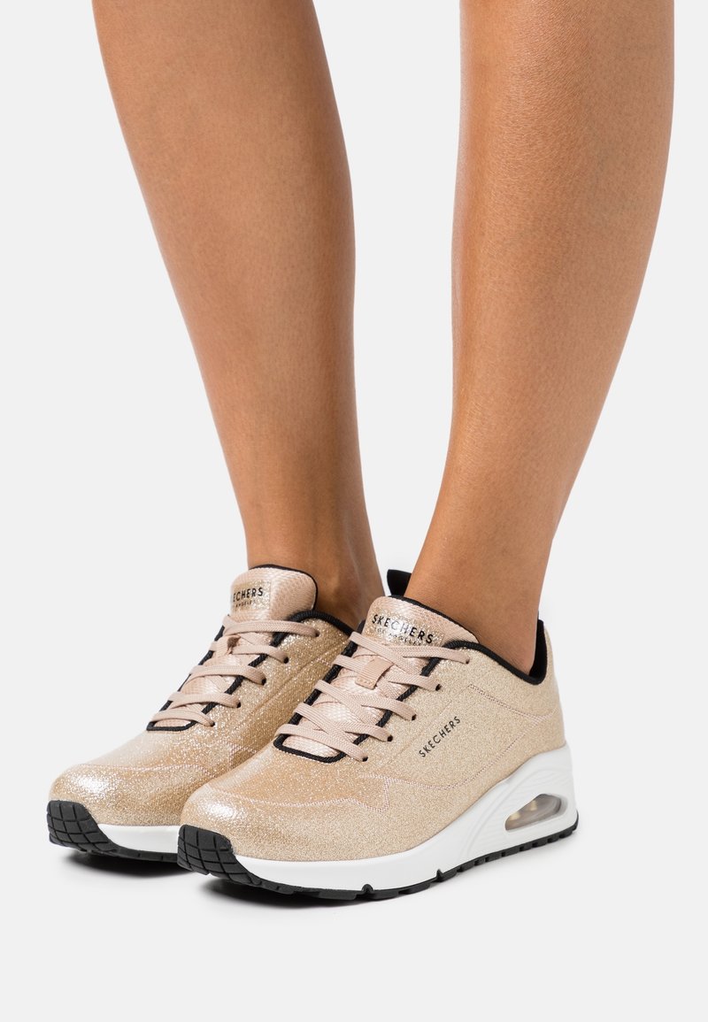 Kvinde iført beige glitrende Skechers sneakers med sort kant, hvide polstrede såler og sort slidbane på hvid baggrund.