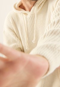 Cremfarbiger, strukturierter Kapuzenpullover mit geripptem Design und großer Kapuze. Verfügt über Zugbänder und einen lockeren Schnitt, wobei ein Teil einer Hand nach vorne greift.