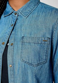 Camisa de mezclilla en azul claro, con collar tipo abotonado, botones en tono dorado, un bolsillo en el pecho y una ligera textura.