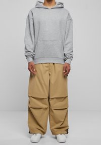 Felpa grigia con tasca frontale, pantaloni beige larghi con dettagli piegati e sneakers bianche, su uno sfondo semplice.