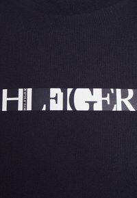 Námornícky bavlněný tričko s bílým grafickým pruhem se stylizovanými písmeny, střídajícími se černými a bílými vzory a kulatým výstřihem.