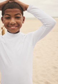 Maglia da surf bianca a maniche lunghe con collo alto, design aderente e texture liscia, indossata da un ragazzo sorridente su una spiaggia.