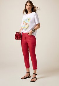 T-shirt blanc avec un graphisme multicolore, associé à un pantalon rouge ajusté très vif et des sandales noires. Les accessoires comprennent une pochette rouge et des bracelets empilés.