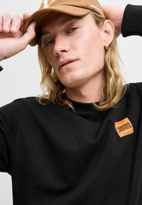 Sudadera negra con cuello redondo, que presenta un logo de Jack & Jones en naranja. La persona lleva una gorra marrón y tiene el cabello largo.