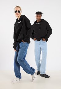 Felpe nere con testo bianco "New York", abbinate a jeans blu e chiari. Sneakers Converse su una, sneakers nere sull'altra.