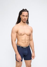 Ανδρικό μοντέλο χωρίς μπλούζα με μακριά dreadlocks, φορούν μπλε ναυτικού χρώματος boxer briefs της Nike, στέκεται μπροστά σε απλό φωτεινό φόντο.