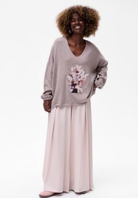 Glückliche Frau mit lockigem Haar trägt einen übergroßen mauvefarbenen Pullover mit floralem Muster, weite hellrosa Hosen und rosa Schuhe und steht vor einem weißen Hintergrund.