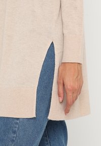 Suéter de punto beige con abertura lateral que muestra unos jeans de mezclilla azules, usado por una persona con la mano descansando cerca de la abertura.