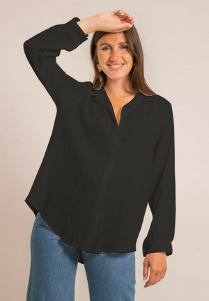 Femme souriante aux longs cheveux bruns, portant un chemisier noir à manches longues et amples et un jean bleu, levant une main au-dessus de sa tête.