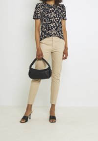 Mustassa letitettävä laukku, jota pitää henkilö, jolla on päällään kukkakuvioinen tummansininen paita ja beiget istuvat housut, jalassa avonaiset mustat korkokengät.