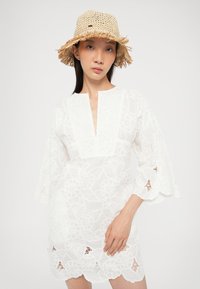 Fehér hímzett tunika V-nyakkivágással és bővülő ujjakkal, virágmintákkal és hullámos szegéllyel. A megfelelője egy szalma balacláva.