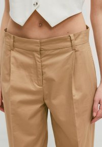 Beige hoge taillebroek met een gladde textuur, voorzien van zijzakken en subtiele plooien aan de voorkant voor een extra ontwerpelement.