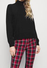 Pull en maille noire avec un col montant et des manches longues, associé à un pantalon à carreaux rouge et noir avec un motif à grille blanche.
