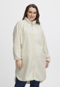 Krämfärgad fleecejacka med hög krage, framtill dragkedja, två sidofickor och mjukt, texturerat tyg. Långa ärmar med uppvullade muddar.
