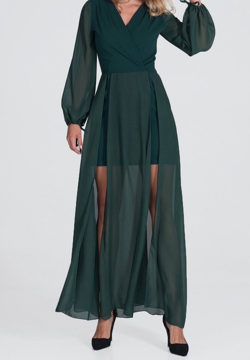 Robe verte cintrée avec des manches longues transparentes et une fente haute sur la cuisse, dotée d'un corsage ajusté et d'une jupe fluide. Des talons noirs complètent l'ensemble.