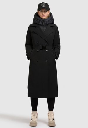 Svart dubbelknäppt trenchcoat med bälte i midjan, lager över en svart puffjacka. Bärs med svarta leggings och ljusbruna sneakers.