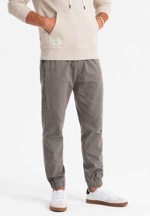 Jogging en coton gris clair avec des poignets élastiques, des poches latérales et des coutures angulaires. Assortis à des baskets blanches avec une semelle marron.