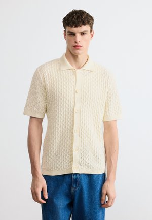 Ung mann iført kremfarget, kortermet strikket skjorte med knapper og blå jeans, stående mot en ensfarget hvit bakgrunn.