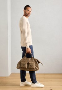 Άντρας σε μπεζ πουλόβερ με πλέξη, ναυτικές παντελόνες και λευκά αθλητικά παπούτσια, κρατώντας μια μεγάλη καφέ δερμάτινη τσάντα duffel, στέκεται μέσα σε έναν εσωτερικό χώρο με ξύλινο δάπεδο.