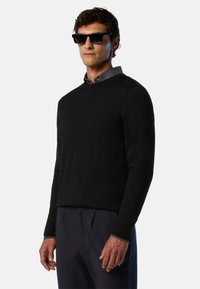 Pull en tricot noir avec un col rond, des manches longues et une texture douce, associé à une chemise grise à col et des lunettes de soleil.