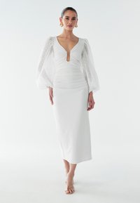 Vit klänning med en figurnära överdel och puffiga, genomskinliga ärmar med prickar. Har en nyckelhålsringning och en slät, förlängd kjol.