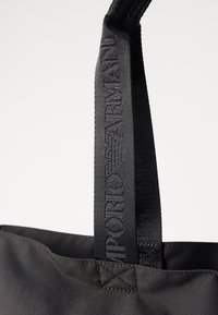 Czarna torba na ramię z teksturowanej tkaniny, z mocnymi uchwytami i wytłoczonym logo na pasku. Prosty, funkcjonalny design.