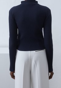 Laivastonsininen ribattu neulepaita, jossa on korkea kaulus, pitkät hihat ja istuva siluetti; yhdistettynä valkoisiin leveälahkeisiin housuihin, joissa on mustia yksityiskohtia.