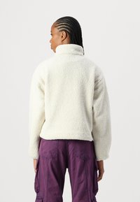 Krémový fleece svetr s vysokým límcem, strukturovaným povrchem a spadlými rameny, spárovaný s purpurovými cargo kalhotami.