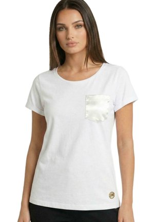 Une femme porte un T-shirt blanc à manches courtes, orné d’une poche poitrine brillante et d’un petit logo doré, sur un fond blanc uni.