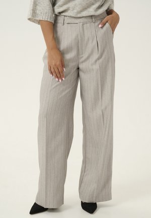 Femme portant un pantalon large gris clair à fines rayures, un pull rentré et des chaussures noires pointues, debout devant un fond uni.