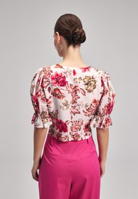 Blumenbluse mit Puffärmeln, die rosa, rote und goldene Muster auf einem hellen Hintergrund zeigt. Kombiniert mit hochgeschnittenen rosa Hosen.