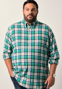 Uomo con barba che indossa una camicia a quadri verde, blu e bianca con maniche arrotolate, in piedi con una mano in tasca e uno sfondo neutro.
