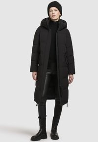 Kvinde står iført en lang sort puffjakke med hætte, sorte læderbukser, sort turtleneck, strikket hat og sorte støvler mod en ensfarvet baggrund.