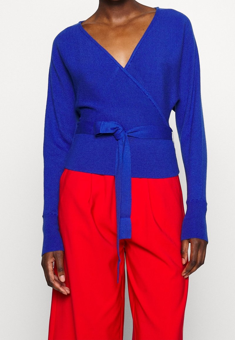 Pull bleu à côtes avec un décolleté en V profond, manches longues et taille nouée. Associé à un pantalon rouge ample pour le contraste.
