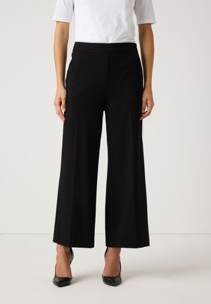 Schwarze weit geschnittene Hosen mit einer glatten Textur, elastischem Bund und Seitentaschen, kombiniert mit einem weißen Kurzarmhemd und schwarzen Absätzen.