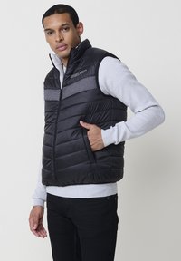 Schwarze gepolsterte Weste mit grauem Horizontalstreifen, Reißverschluss und vorderen Taschen. Getragen über einem hellgrauen strukturierten Pullover, mit hohem Kragen.