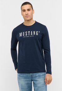 Granatowa, długorękawowa koszulka T-shirt z białym logo "MUSTANG" na piersi, wykonana z miękkiego materiału, zestawiona z wyblakłymi niebieskimi dżinsami.